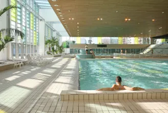 Indoor municipal swimming pool at Grand Parc in Bordeaux featuring an Olympic-sized pool, swimmers, lounge chairs, palm trees, and modern glass architecture.