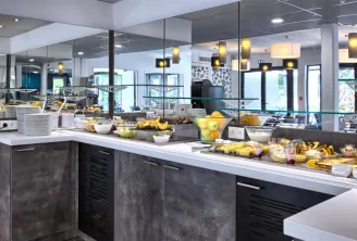 Modern breakfast buffet with black and white counter, fresh fruits, pastries and beverages in a bright room with mirrors and designer lighting at Hôtel Au Comté d'Ornon in Bordeaux.