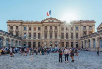 Facade of Palais Rohan in Bordeaux with its paved courtyard, classical stone architecture, French flag on top, and visitors in the courtyard on a sunny day.