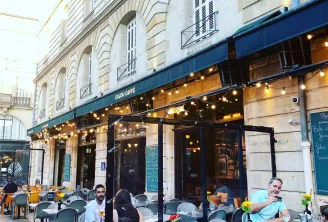 Outdoor seating area of Cajou Caffé café in Bordeaux with customers at tables, Haussmann-style building facade and blue awning.