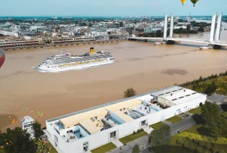 Aerial view of Bordeaux showing the Garonne River with a cruise ship, colorful hot air balloons flying over La Fabrique Pola, a modern white waterfront building, the Chaban-Delmas Bridge in the background, and the city skyline.