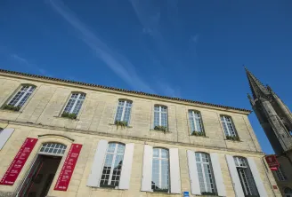 Facade of the Maison du Vin de Saint-Emilion in Bordeaux, a stone building with multi-pane windows, red information banners and a Gothic bell tower visible on the right