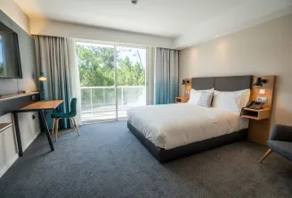 Modern hotel room at Holiday Inn Bordeaux Mérignac featuring a queen-size bed, work desk, wall-mounted TV, and glass balcony overlooking trees