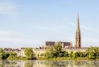 Panoramic view of the Basilica of Saint-Michel in Bordeaux with its Gothic spire towering over the urban landscape, surrounded by Haussmann-style buildings and reflected in the Garonne river waters.