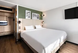 Modern hotel room featuring a double bed with white linens, beige headboard, green accent wall, wall-mounted TV, light wood wardrobe and bunk beds in the background