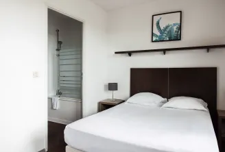 Modern bedroom with double bed with white linens, black headboard, adjoining bathroom with glass shower, minimalist décor and botanical artwork.