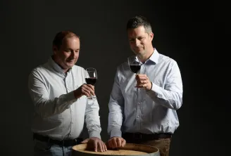 Two men in white shirts toasting with glasses of red wine in front of a wooden barrel in the cellars of Château Lagrange in Bordeaux.