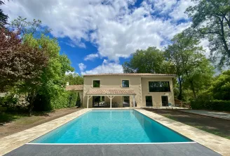 Modern villa with rectangular swimming pool surrounded by lawn and trees, beige facade with covered terrace, cloudy blue sky.