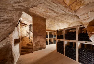 Underground wine cellar of the Jacobins Convent in Bordeaux featuring rows of wine bottles stored in wooden racks, exposed stone walls, heavy wooden door, and a metal cross.