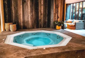 Indoor spa featuring a turquoise hot tub with massage jets, surrounded by rustic wooden walls, woven baskets, and warm-toned relaxation furniture.