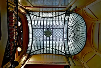 Overhead view of the stained glass ceiling at Hôtel Frugès in Bordeaux, displaying an ornate geometric structure with decorative patterns, gilded wooden frame, and Art Deco lighting fixtures.