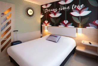 Modern hotel room featuring a white double bed, illuminated headboard, accent wall with red and white floral patterns, circular golden wall decoration, and inspirational text « Dreams come true »