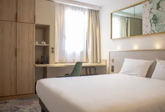 Modern hotel room featuring a double bed with white linens, integrated desk, light wood wardrobe, gold-framed mirror and window with beige curtains in Bordeaux