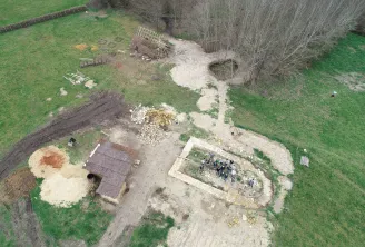 Aerial view of a medieval archaeological excavation site showing dig areas, stone foundations, construction materials, and archaeologists working in a grassy field bordered by trees.