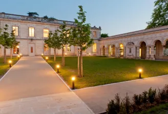 Illuminated courtyard of Table de La Chèze in Bordeaux, featuring stone buildings, manicured lawn, paved walkways and evening ambient lighting