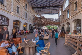 Interior view of Darwin, Bordeaux's covered public space featuring restaurants, bars and shops. The pedestrian area hosts visitors seated at tables and chairs, with brick architecture, arcades, and an open metal roof.