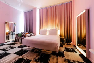 Modern bedroom at Mama Shelter Bordeaux featuring a white bed, soft pink walls, gradient orange and gold curtains, illuminated mirrors, striped black and white armchair, and geometric black and beige carpet