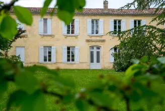 Facade of Château Valoux in Bordeaux, a beige stone manor with blue shutters, surrounded by green lawn and vegetation.
