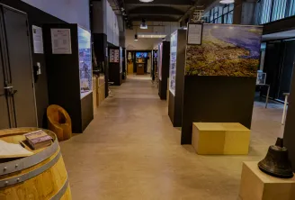 Interior of the Bordeaux Maritime History Museum displaying an exhibition gallery with informational panels, wooden barrels, display cases and historical maritime objects, lit by suspended lamps.
