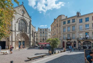 Sunny square in Bordeaux featuring the Saint-Pierre Church with its ornate Gothic facade and clock, surrounded by Haussmann-style stone buildings, with visitors and café terraces.
