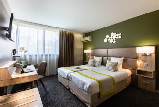 Modern hotel room featuring two twin beds, green walls, wooden desk with chair, large windows with beige curtains, white circular wall decoration, and light wood furniture.