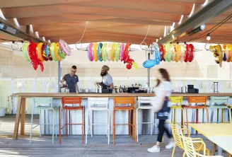Modern bar at Mama Shelter in Bordeaux featuring wooden counter, colorful bar stools, staff members working, and festive hanging bracelet decorations on the ceiling.
