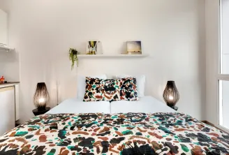 Modern bedroom featuring a double bed with colorful geometric patterned sheets, two designer bedside lamps, white pillows, wall shelf with decor, kitchenette on the left and window on the right