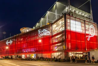 The UCPA Sport Station in Bordeaux lit up at night, a modern building with red façade and glass panels, surrounded by visitors and parked bicycles.