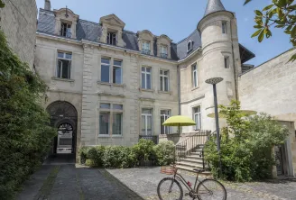 Elegant facade of a Bordeaux mansion hotel with round tower, paved courtyard, parked bicycle, yellow parasol and greenery.