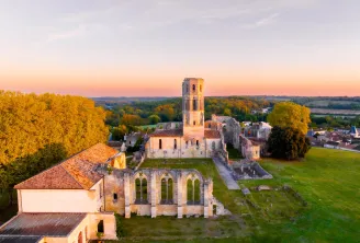 Aerial view at sunset of La Sauve-Majeure Abbey near Bordeaux, showing the church ruins with its Romanesque tower and adjacent buildings surrounded by green fields and forests.
