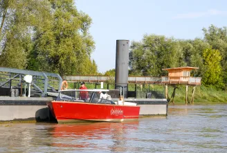 The red Grattequina water taxi carries passengers on the Garonne River in Bordeaux, passing by port facilities and riverside vegetation.