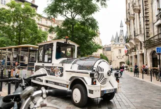 A white and black electric tourist train traveling down a cobblestone street in Bordeaux, with Haussmann-style buildings and a castle in the background.