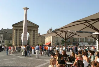 Victory Square in Bordeaux featuring the commemorative column, a church, outdoor café umbrellas, and crowds of visitors on a sunny day.