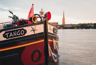 The Tango boat moored on the Garonne River in Bordeaux, decorated with vibrant colors, a red flag, musical instruments and ornaments, with the Saint-Michel Basilica visible in the background.
