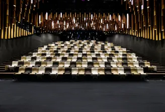 Interior of the Thomas Jefferson Auditorium at La Cité du Vin in Bordeaux, featuring tiered seating in beige and black tones, with an illuminated ceiling featuring golden and white light panels.