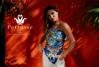 Woman wearing a blue strapless top with gold and white geometric patterns, posing against a red background with Petrusse Paris logo, French maison, with green plants in the background.