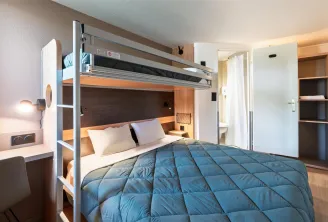 Modern hotel room featuring a metal and white bunk bed with blue quilt, white bedding, integrated desk with lamp, white wardrobe, visible bathroom, light wooden flooring and warm ambient lighting.