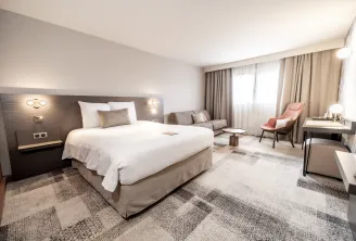 Modern hotel room at Mercure Bordeaux-Lac featuring a king-size bed, grey upholstered headboard with wall sconces, gray sofa, pink accent chair, large windows with beige curtains, contemporary furnishings, and neutral color scheme.