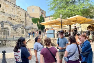 A group of tourists listening to a tour guide in front of a historic church in Bordeaux, sheltered under yellow umbrellas, with stone buildings and green trees in the background
