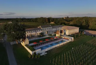 Aerial view of Château Fage, a luxury hotel-restaurant with ochre stone buildings, blue swimming pool, red loungers on terrace, surrounded by vineyards and forests in the Bordeaux region.