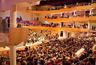 Interior view of the Bordeaux Auditorium featuring an orchestra performing on stage with musicians and instruments, surrounded by spectators seated in multi-level tiered seating. Modern architecture with light wood balconies and purple and orange ambient 