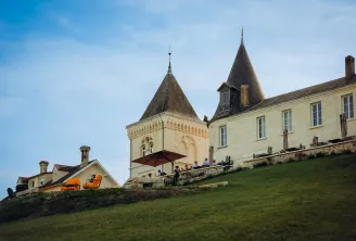 CarlMagnus Castle near Bordeaux, Roux Oulié house featuring distinctive conical towers, pale yellow facade, shaded terrace with orange chairs overlooking green grounds.