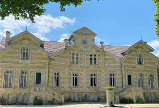 Front view of Château Maucaillou in Bordeaux, a grand pale yellow stone mansion with red tile roof, symmetrical pavilions, set on manicured lawn with garden furniture in the foreground.