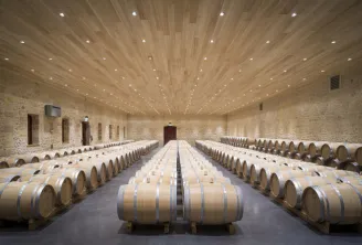 Wine aging cellar at Château Fourcas Hosten in Bordeaux, featuring hundreds of wooden barrels arranged symmetrically beneath a light wood ceiling with integrated lighting