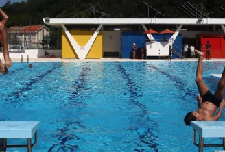 Municipal swimming pool in Floirac, Bordeaux, with multiple swimmers and divers enjoying the turquoise water, colorful buildings visible in the background