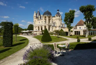 White Belle Époque mansion with black domes surrounded by manicured gardens, lounge chairs in foreground and shaded terrace in background
