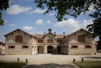 Symmetrical main building of the Horse Museum in Bordeaux, featuring classical architecture with beige stone facade, red tile roof and red wooden doors, set in a paved courtyard.