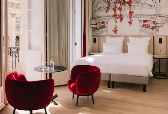 Elegant bedroom at Hôtel Majestic in Bordeaux featuring a white bed, two burgundy velvet armchairs, a balcony overlooking classical Bordeaux architecture, and red floral wall decor.