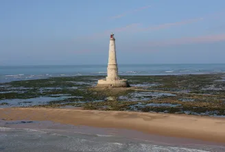 Cordouan lighthouse, a white and beige lighthouse with a red dome, standing on a rocky reef covered with seaweed off the coast of Bordeaux, surrounded by sandy beaches and the Atlantic Ocean.