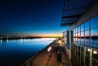 Cap Sciences building at sunset in Bordeaux, with glass facades reflecting the orange sky, overlooking the Garonne River. A lit pedestrian walkway runs along the waterfront.
